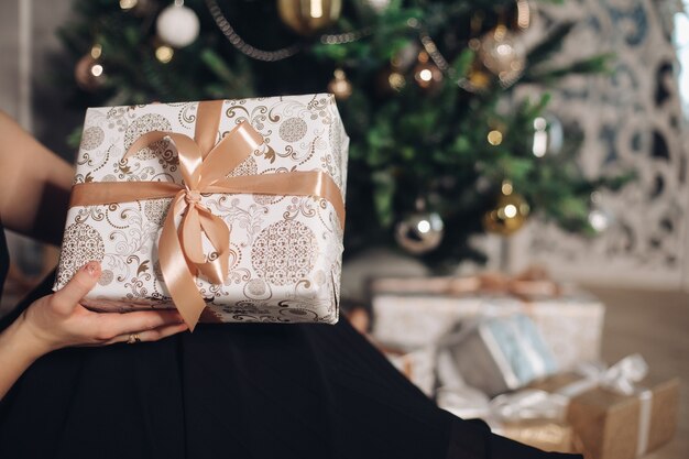 Una pequeña caja con un regalo de Año Nuevo en la mano frente al árbol de Navidad