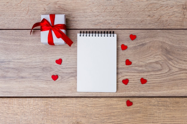 Pequeña caja de regalo con bloc de notas y corazones rojos en mesa