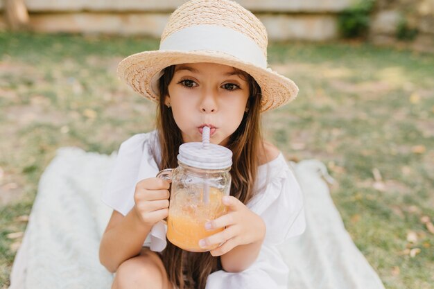 Pequeña dama pensativa con sombrero de verano con cinta blanca bebe jugo de naranja y mirando a otro lado. retrato al aire libre de una niña de cabello castaño disfrutando de un cóctel en una manta en el parque.