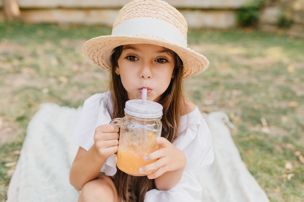 Pequeña dama pensativa con sombrero de verano con cinta blanca bebe jugo de naranja y mirando a otro lado. Retrato al aire libre de una niña de cabello castaño disfrutando de un cóctel en una manta en el parque.