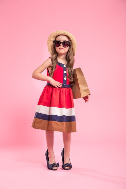 pequeña fashionista con una bolsa de compras en un sombrero de verano y gafas, sobre un fondo de color rosa en los zapatos de mamá, el concepto de moda infantil