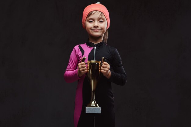 La pequeña y feliz patinadora campeona sostiene la copa de un ganador. Aislado en un fondo de textura oscura.