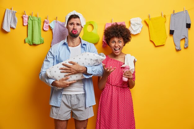 La pequeña hija recién nacida duerme cómodamente en el abrazo de los padres. Pareja cansada se preocupa por el bebé. Papá molesto con pañal en la cabeza, sostiene al bebé envuelto con una manta. Madre irritada va a alimentar a un niño