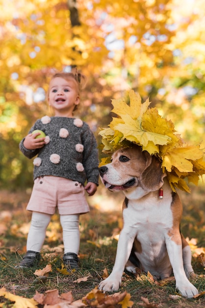 Pequeña muchacha que se coloca cerca del perro del beagle que lleva el sombrero de la hoja del otoño en bosque