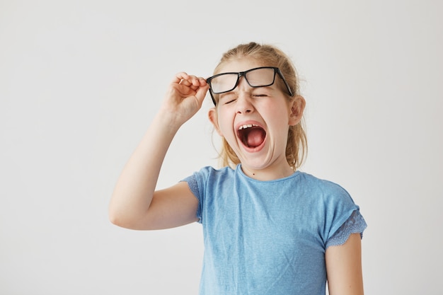 Pequeña muchacha rubia divertida con los ojos azules bonitos que levantan los vidrios y que bostezan con los ojos cerrados en clase porque la lección aburrida larga.