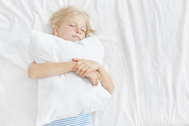 Pequeña niña adorable con cabello rubio, cara pecosa, cerrando los ojos, abrazando la almohada blanca, durmiendo agradablemente en la ropa de cama blanca. Niño que tiene sueños agradables en la mañana descansando en casa