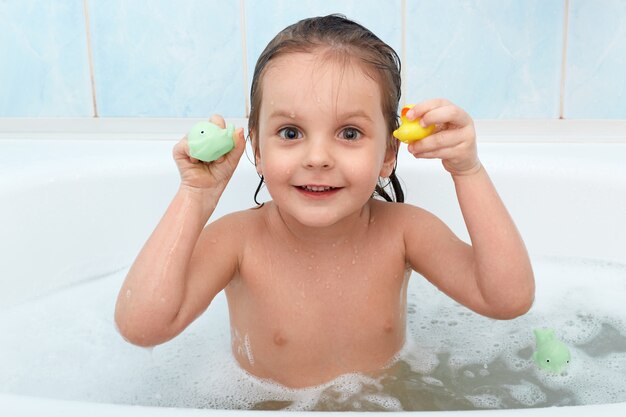 Pequeña niña con juguete en manos tomando baño, juega en el agua con pato y delfín