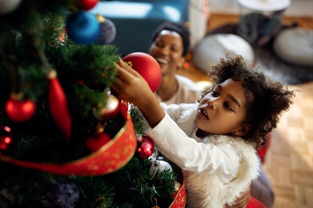 Pequeña niña negra decorando el árbol de Navidad en casa