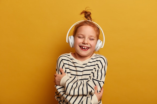 La pequeña niña pelirroja se abraza, se ríe y se divierte, escucha música en auriculares estéreo, usa un jersey de rayas, posa sobre una pared amarilla, pasa el tiempo libre en su pasatiempo favorito, se divierte