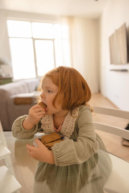 La pequeña niña pelirroja caucásica usa un vestido que disfruta de deliciosas galletas mientras se sienta a la mesa en la sala de luz Concepto de niños y nutrición