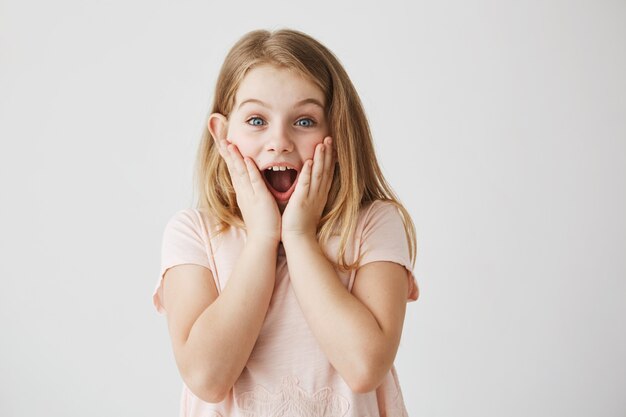 Pequeña niña rubia con ojos azules, sosteniendo la cara con las manos y la boca abierta expresando emoción. Niño feliz después de recibir el regalo de cumpleaños de la madre.