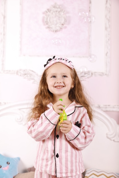 Una pequeña pelirroja vestida con pijama rosa está jugando con burbujas en la habitación en la cama grande