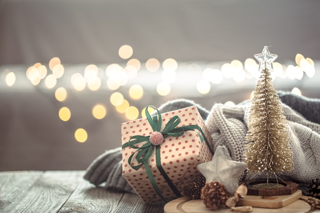 Pequeño árbol de Navidad con presente sobre luces de Navidad bokeh en casa sobre mesa de madera con suéter en una pared y adornos.