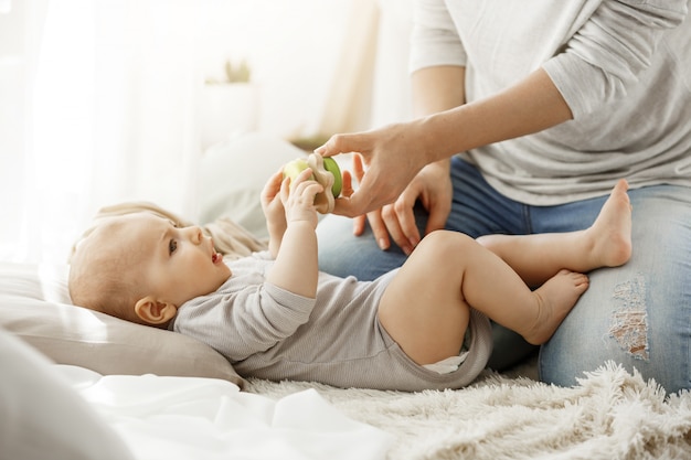 Pequeño bebé que pasa infancia feliz con la madre joven. Niño tratando de tomar un hermoso juguete de tiernas manos de mamá. Concepto de familia