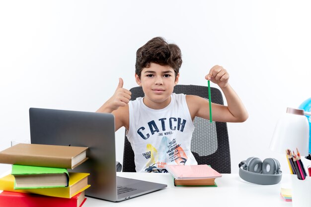 Pequeño colegial complacido sentado en un escritorio con herramientas escolares sosteniendo un lápiz con el pulgar hacia arriba aislado sobre fondo blanco