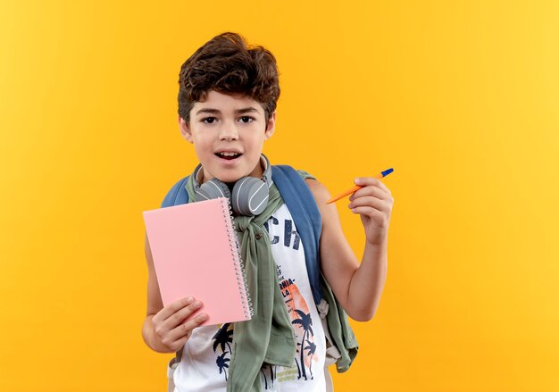 Pequeño colegial complacido vistiendo bolsa trasera y auriculares sosteniendo el cuaderno con bolígrafo