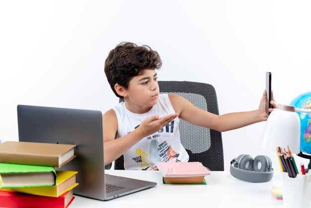 Pequeño colegial confundido sentado en un escritorio con herramientas escolares sosteniendo y mirando el teléfono