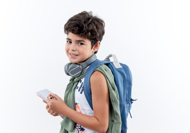 Pequeño colegial contento con bolsa trasera y auriculares con cuaderno y bolígrafo aislado sobre fondo blanco.