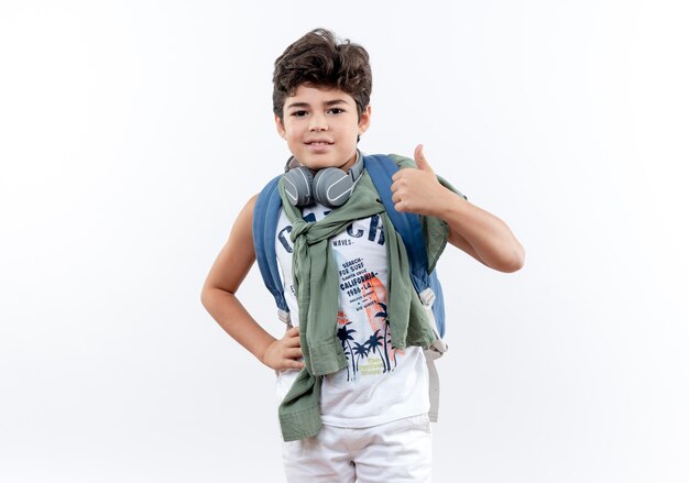 Pequeño colegial contento con bolsa trasera y auriculares poniendo la mano en la cadera con el pulgar hacia arriba