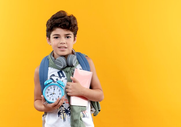 Pequeño colegial contento con bolsa trasera y auriculares sosteniendo libro con reloj despertador aislado sobre fondo amarillo con espacio de copia