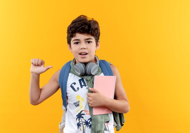 Pequeño colegial contento con bolsa trasera y auriculares sosteniendo portátil aislado sobre fondo amarillo