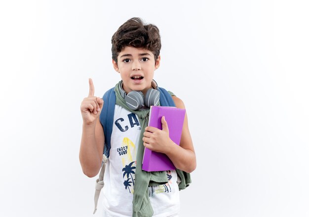 Pequeño colegial impresionado con mochila y auriculares sosteniendo el libro y apunta hacia arriba aislado sobre fondo blanco con espacio de copia