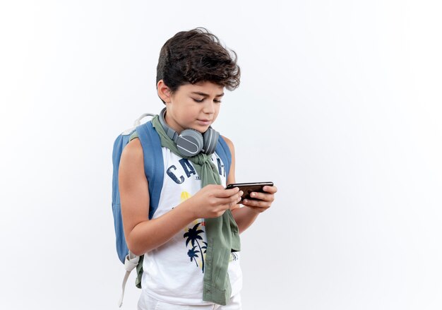 Pequeño colegial con mochila y auriculares jugando en el teléfono