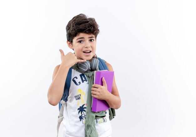 pequeño colegial con mochila y auriculares sosteniendo un libro y mostrando un gesto de llamada telefónica