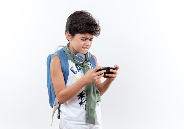 Foto gratuita pequeño colegial preocupado con mochila y auriculares jugando en el teléfono