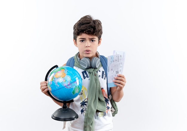 Foto gratuita pequeño colegial preocupado vistiendo bolsa trasera y auriculares sosteniendo boletos y globo aislado sobre fondo blanco.