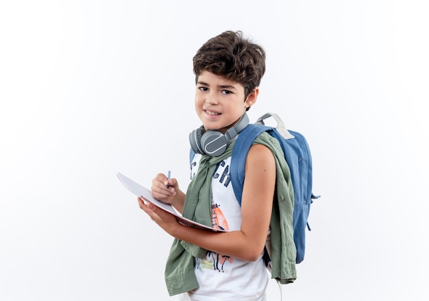 Pequeño colegial satisfecho con bolsa trasera y auriculares sosteniendo el cuaderno con lápiz aislado sobre fondo blanco con espacio de copia