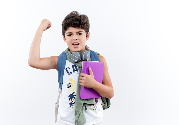 Pequeño colegial seguro con mochila y auriculares sosteniendo el libro y haciendo un gesto fuerte