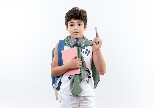 Pequeño colegial sorprendido con bolsa trasera y auriculares sosteniendo el cuaderno y levantando la pluma aislada en blanco