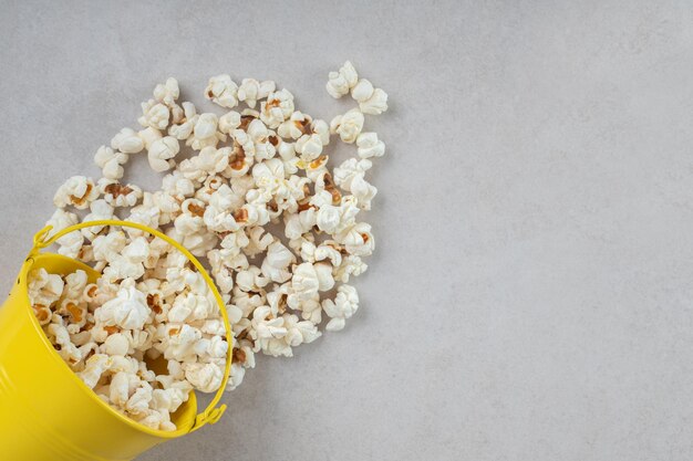 Pequeño cubo amarillo derramando palomitas de maíz frescas sobre la mesa de mármol.