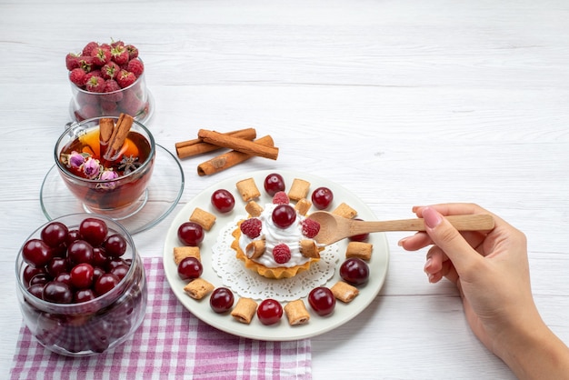 Foto gratuita pequeño y delicioso pastel con frambuesas, cerezas y galletas pequeñas, té, canela en un escritorio de luz, té con crema de frutas y bayas