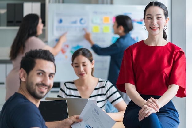 Pequeño emprendedor asiático inteligente de pie retrato confiado con amigos socios reunión informal lluvia de ideas con pizarra blanca y análisis de gráficos de papel de finanzas comerciales en el fondo de la oficina