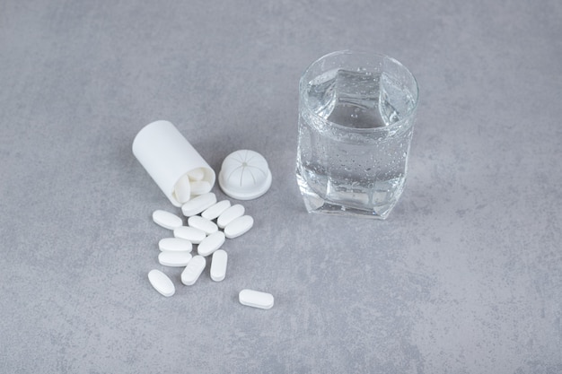 Un pequeño frasco blanco de pastillas blancas con un vaso de agua pura sobre una superficie gris