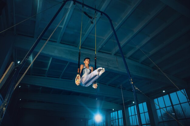 Pequeño gimnasta masculino entrenando en gimnasio, flexible y activo. Chico caucásico en forma, atleta en ropa deportiva blanca practicando ejercicios de equilibrio en los anillos. Movimiento, acción, movimiento, concepto dinámico.