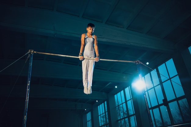 Pequeño gimnasta masculino entrenando en gimnasio, flexible y activo. Niño caucásico, atleta en ropa deportiva blanca practicando ejercicios de fuerza, equilibrio. Movimiento, acción, movimiento, concepto dinámico.