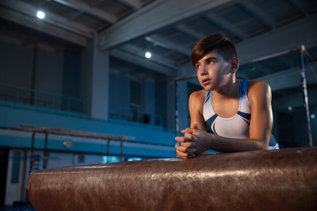 Pequeño gimnasta masculino entrenando en gimnasio, flexible y activo. Niño pequeño caucásico, atleta en ropa deportiva blanca que publica seguro.