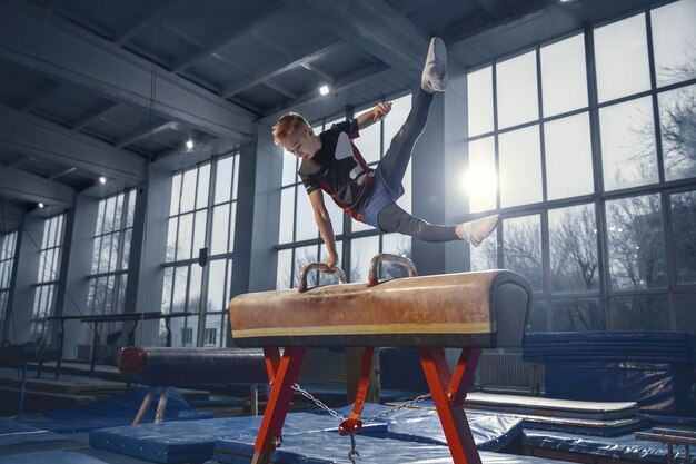 Pequeño gimnasta masculino entrenando en gimnasio, flexible y activo. Niño pequeño caucásico, atleta en ropa deportiva practicando ejercicios de fuerza, equilibrio. Movimiento, acción, movimiento, concepto dinámico.