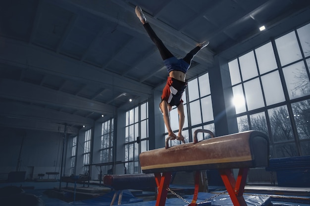 Pequeño gimnasta masculino entrenando en gimnasio, flexible y activo. Niño pequeño caucásico, atleta en ropa deportiva practicando ejercicios de fuerza, equilibrio. Movimiento, acción, movimiento, concepto dinámico.