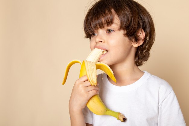 Pequeño kd adorable adorable dulce comiendo plátano en camiseta blanca sobre escritorio rosa