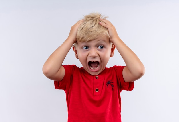 Un pequeño y lindo niño rubio sorprendido en camiseta roja sosteniendo la cabeza con las manos mientras mira de lado en una pared blanca