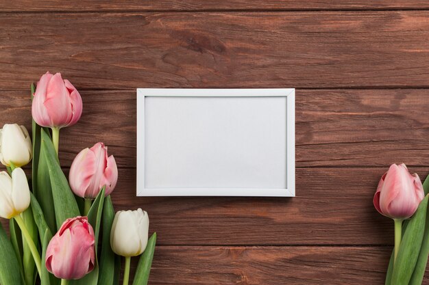 Pequeño marco en blanco blanco con tulipanes rosados y blancos en el escritorio de madera