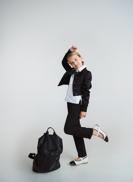 Pequeño modelo caucásico femenino posando en uniforme escolar con mochila sobre fondo blanco.