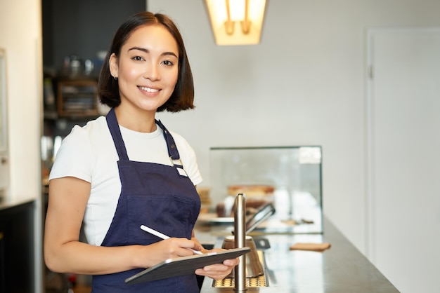 Pequeño negocio de cafetería sonriente chica asiática barista en delantal usando tableta como café de procesamiento de terminal pos