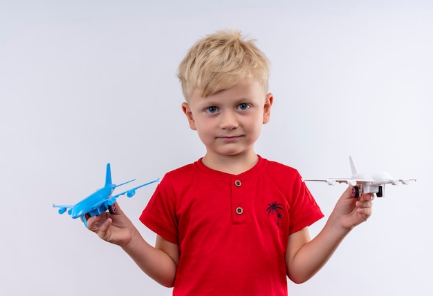 Un pequeño niño lindo con cabello rubio y ojos azules con camiseta roja volando avión de juguete azul y blanco mientras mira en una pared blanca