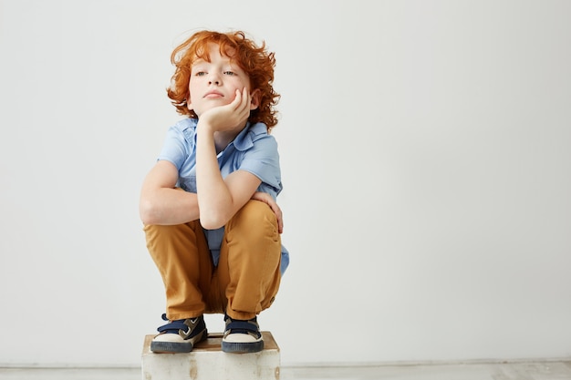 Pequeño niño pelirrojo divertido sentado, sosteniendo la cabeza con la mano