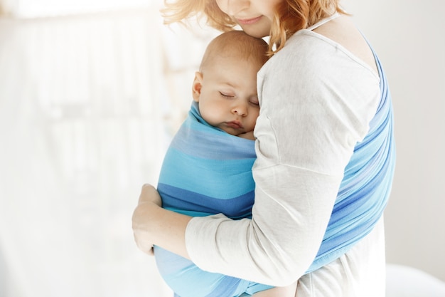 El pequeño niño recién nacido cierra los ojos y duerme bien en la honda del bebé sintiéndose protegido de su hermosa joven madre. Familia, concepto de estilo de vida.